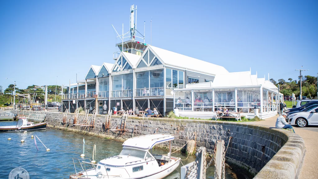 The exterior of Mornington foreshore restaurant, The Rocks, a clear, sunny day, with a small white boat in the harbour below.