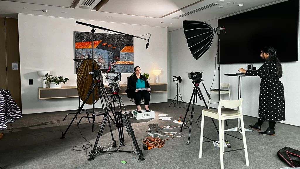 The set of a corporate film in a Melbourne building, featuring 3 lights, 3 cameras, a standing interviewer on the right hand side of frame, and a seated interviewee central.