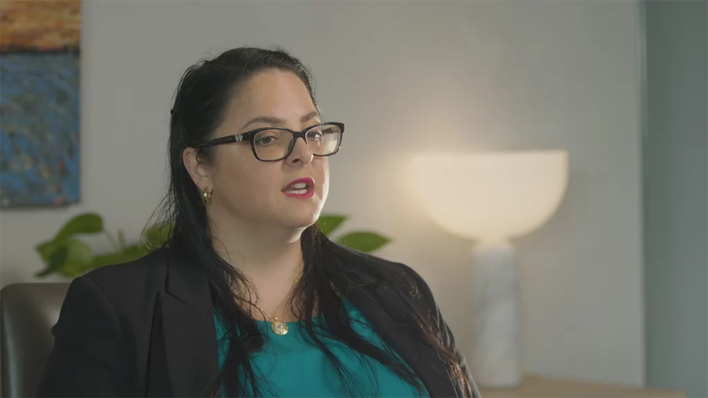 A larger middle aged, caucasian woman with red lip stick, glasses and black long hair being interviewed.