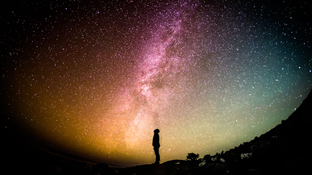 A man silhouetted at night time to the backdrop of stars and a colourful sky.