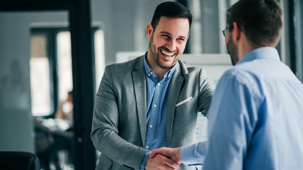 Two male businessmen shake hands next to a window in an office.