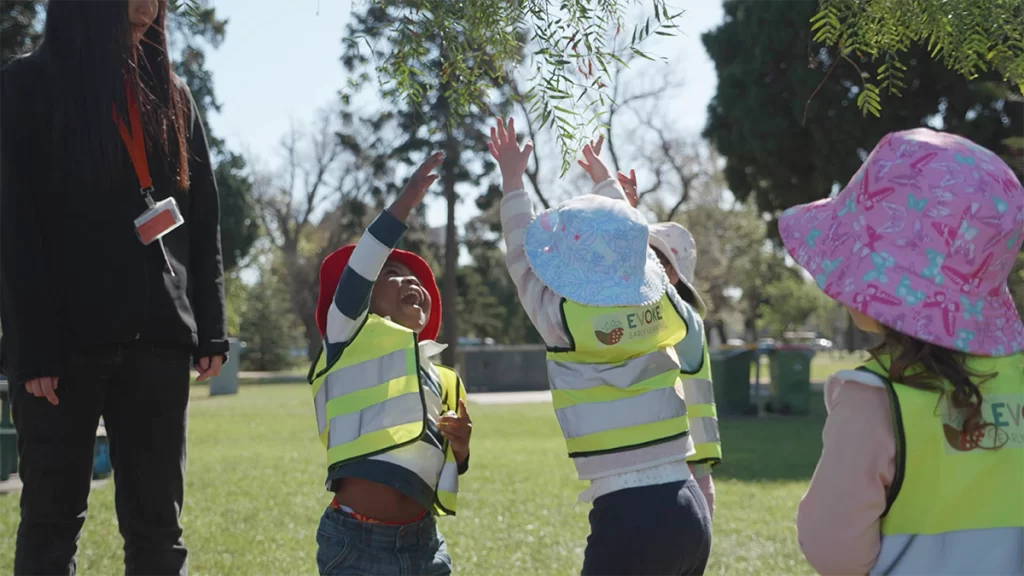 Two children from Evoke Early Learning jumping to try to reach an overhanging branch of small leaves.