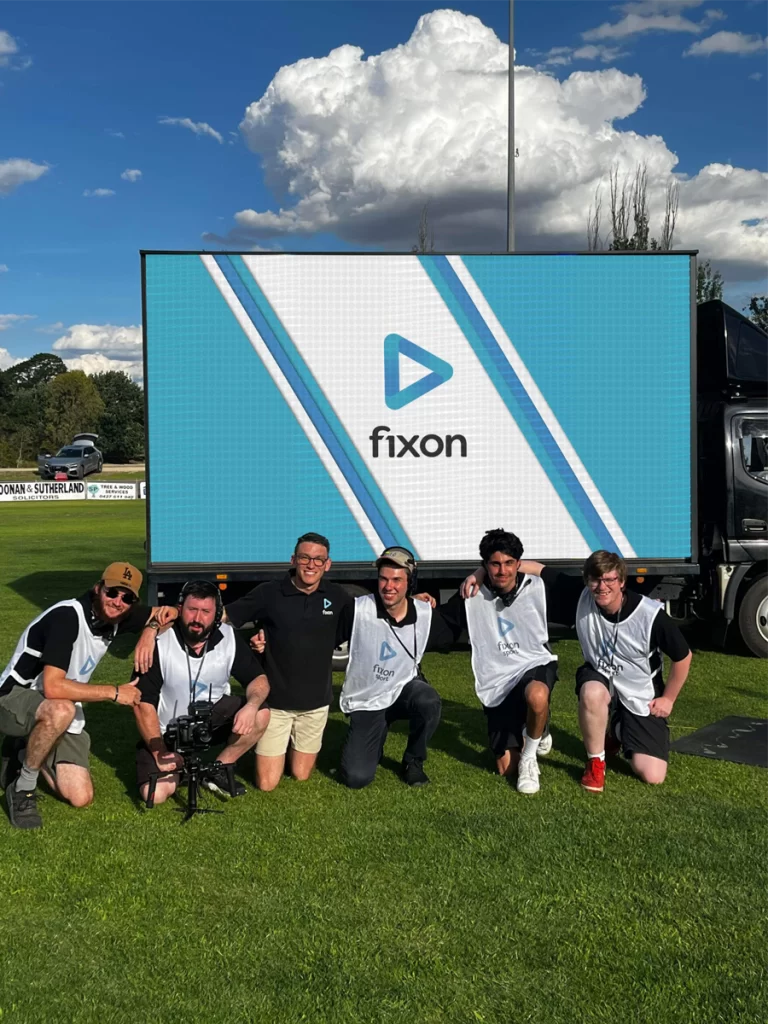 6 Fixon Media Group kneeling in a line on grass. Their arms around each other's shoulders, with a Fixon Media Group logo being displayed on a large digital screen behind them.