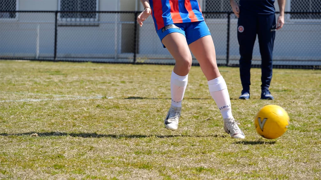 A young female player from Mount Martha Soccer Club kicks a yellow soccer ball on a grass pitch.