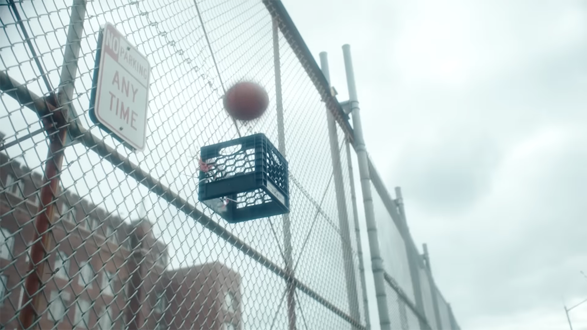 A basketball hits a caged fence, as it looks to go through a makeshift basketball hoop made out of a crate.