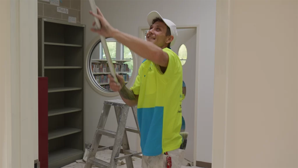 A painter using a long armed roller to paint an interior wall white.