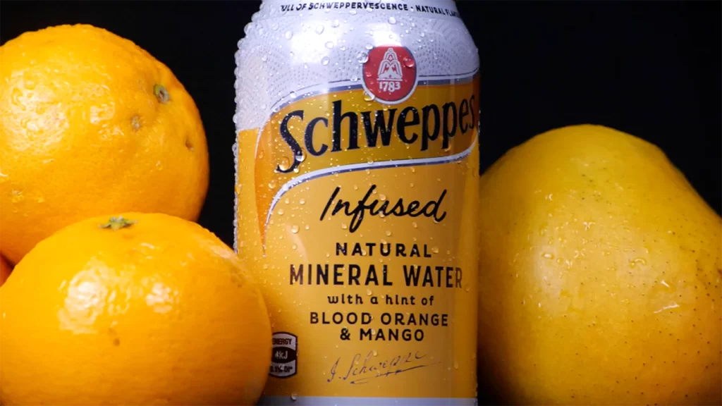 2 oranges and a mango accompany either side of a Schweppes can of Blood Orange and Mango mineral water.