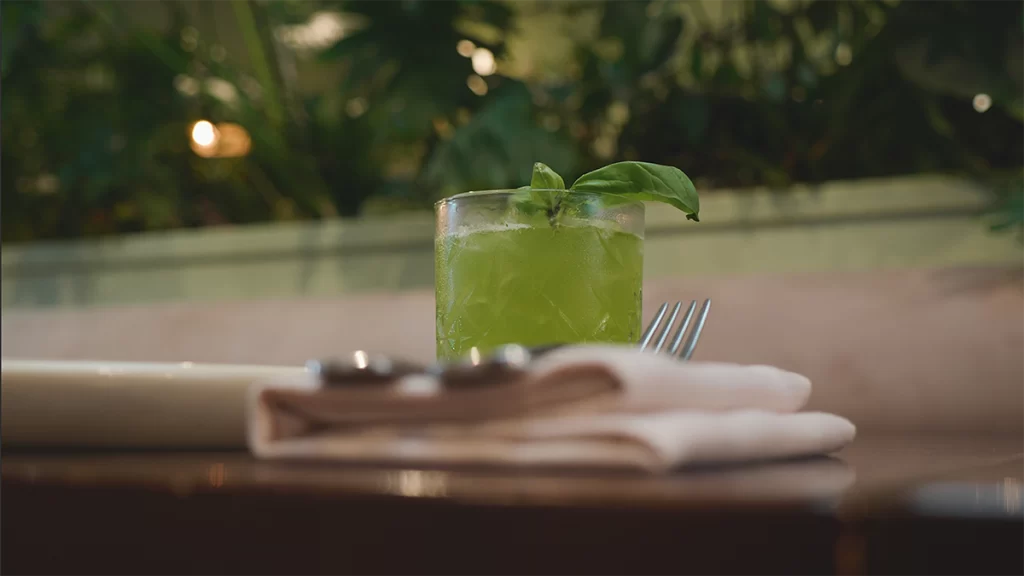 A green coloured cocktail with a handful of basil leaves overhanging the right hand side of a rock glass.