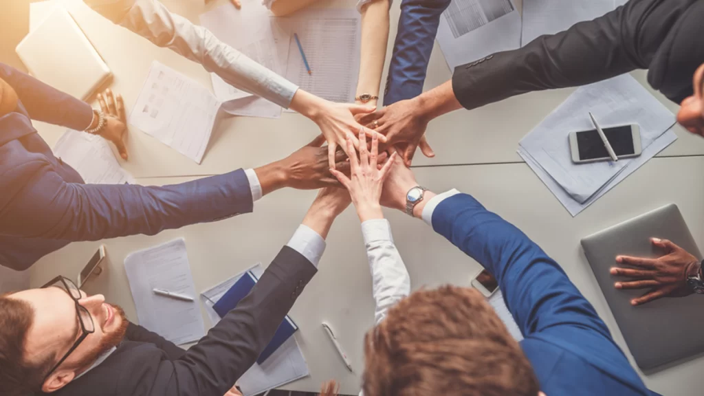 8 office workers of mixed genders and ethnicities place their hands on top of each other in the middle of a circle.