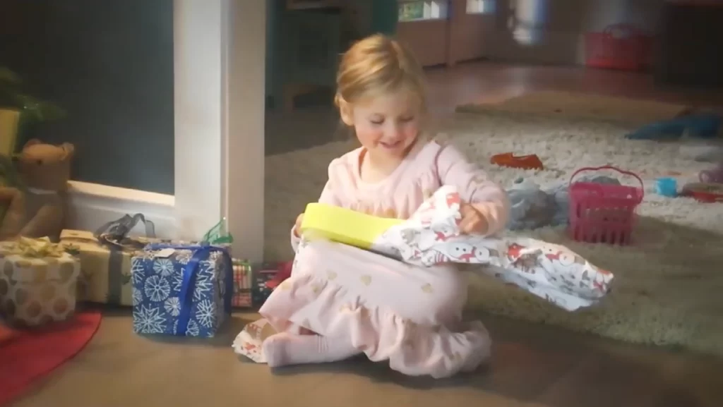 A young girl opens a small guitar under a Christmas tree.